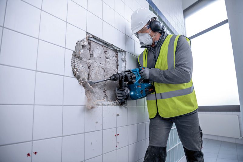 Person wearing safety equipment uses a cordless rotary hammer to break through a tiled wall.