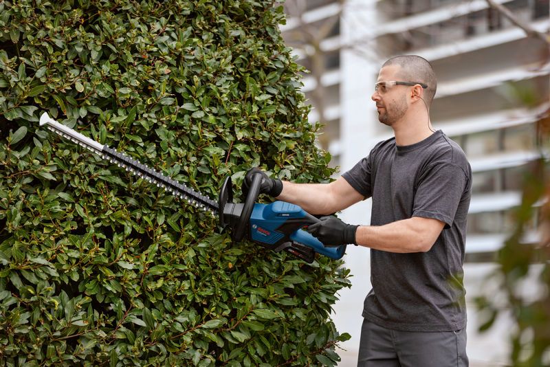 Person wearing safety equipment trims a bush outdoors using a cordless hedge trimmer.