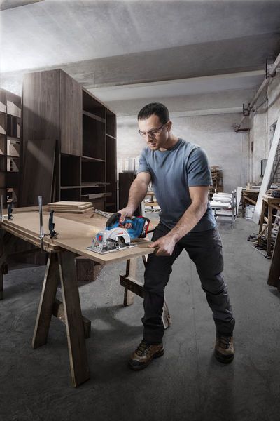 A person wearing safety equipment cuts wood with a cordless circular saw in a workshop.