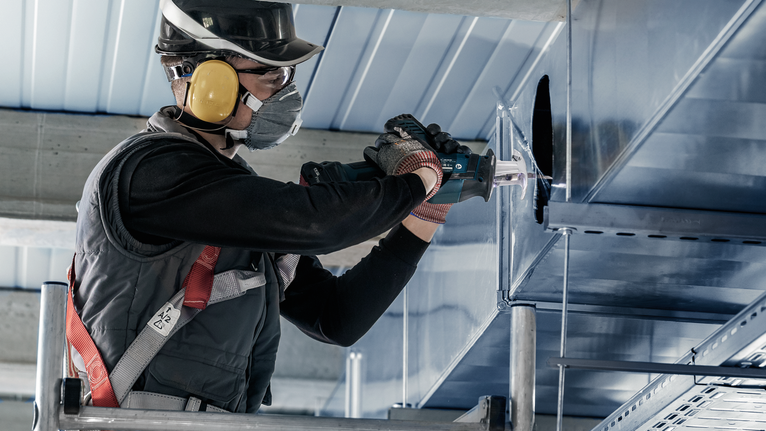 A person wearing safety equipment drills into a metal ventilation duct.