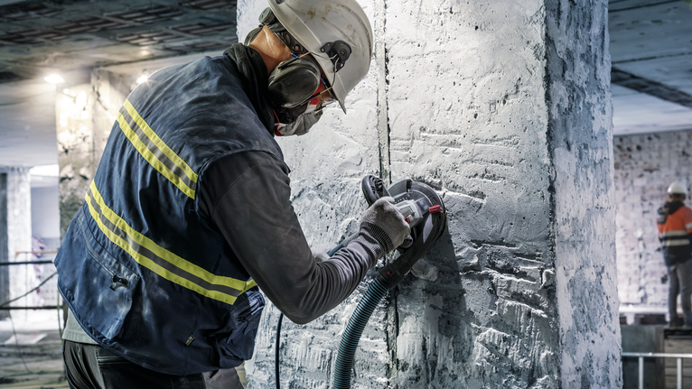 Person wearing safety equipment grinds a concrete pillar with a power tool.