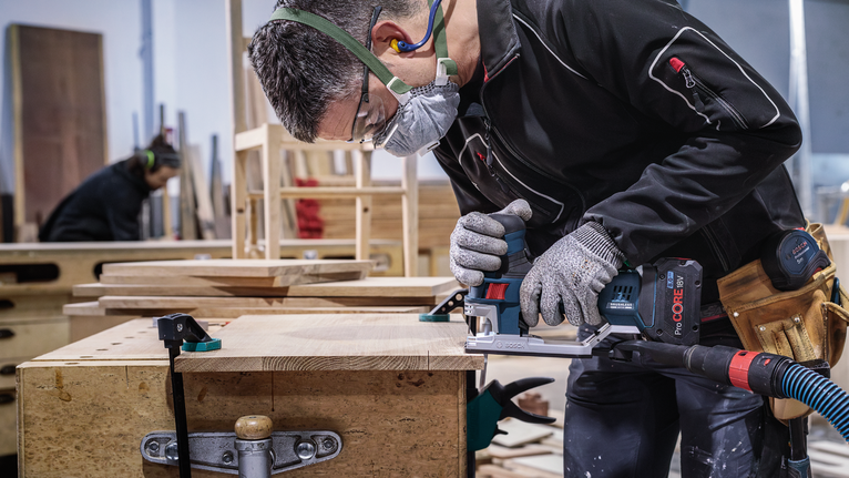 A person wearing safety equipment uses a power tool to cut wood on a workbench.