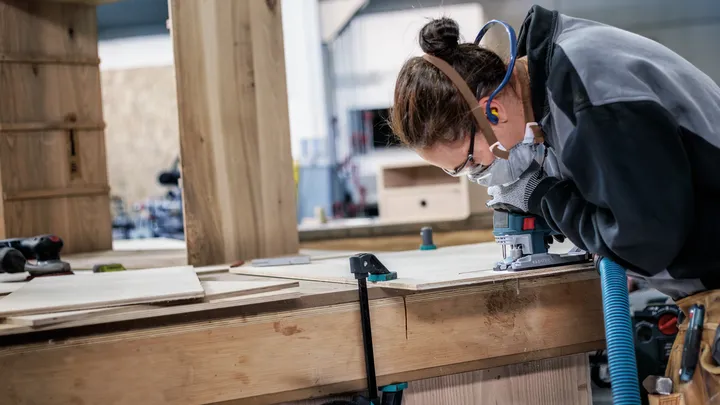 A person wearing safety equipment uses a woodworking tool on a wooden board.