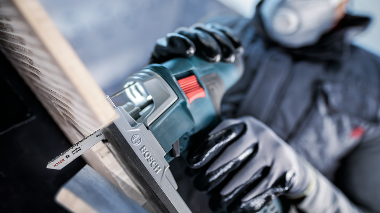 Person wearing safety equipment uses a reciprocating saw to cut a wooden board.