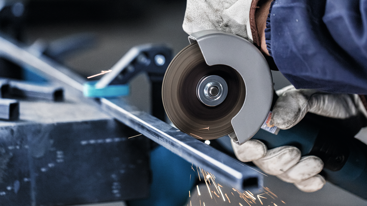 A worker wearing safety equipment cuts a metal bar with an angle grinder.