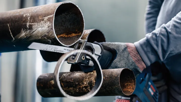 A person wearing safety equipment cuts through a metal pipe with a reciprocating saw.
