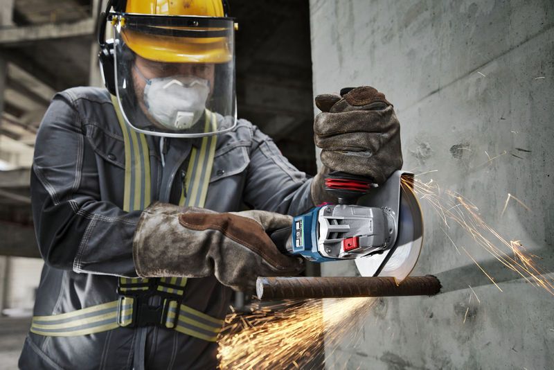 A person wearing safety equipment uses a cordless angle grinder to cut through rebar.