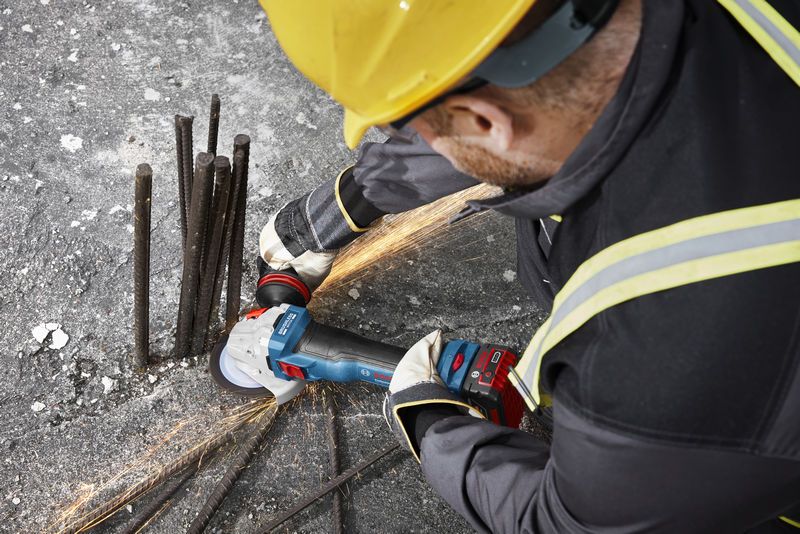 A person wearing safety equipment grinds rebar on concrete with a cordless angle grinder.