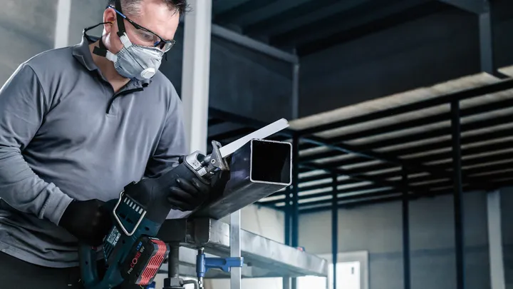 Person wearing safety equipment cuts a metal beam with a reciprocating saw.