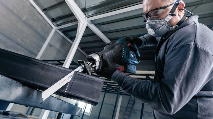 Person wearing safety equipment cuts a metal beam with a reciprocating saw.