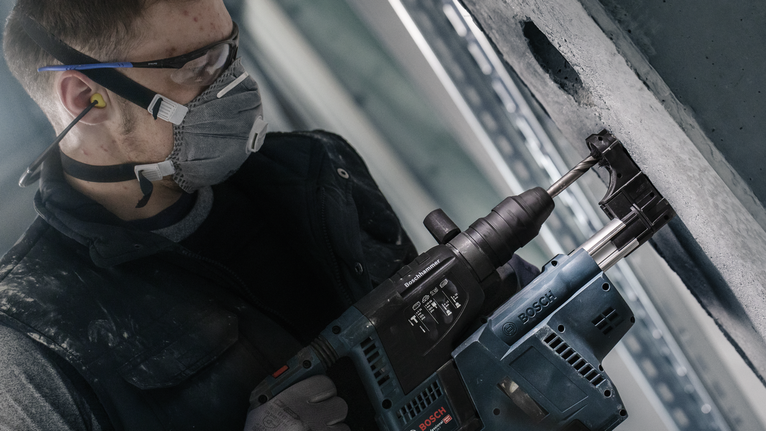 A person wearing safety equipment drills into a concrete wall with a power tool.
