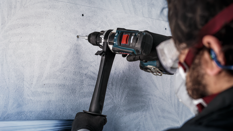 A person wearing safety equipment drills into a concrete wall.