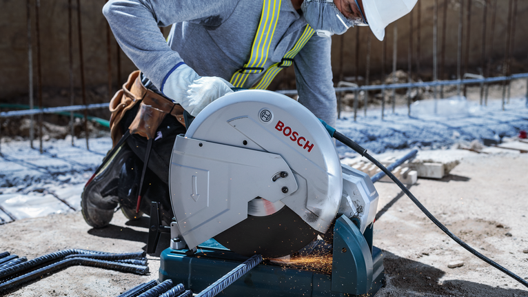 Worker wearing safety equipment cuts rebar with a metal chop saw, creating sparks.