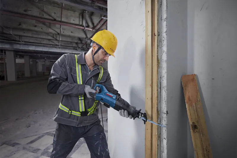 A person wearing safety equipment cuts through a wall stud with a reciprocating saw.