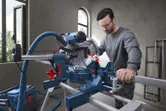 A person wearing safety equipment cuts wood on a mitre saw in a workshop.