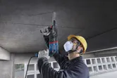A person wearing safety equipment drills into a concrete ceiling using a rotary hammer.
