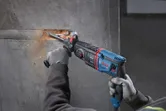 A person wearing safety equipment uses a rotary hammer to chisel a groove in concrete.