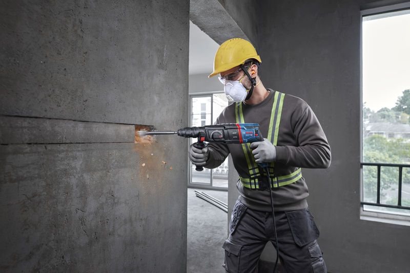 A person wearing safety equipment uses a rotary hammer to drill into a concrete wall.