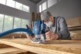A person wearing safety equipment uses a router on a wooden plank in a workshop.