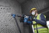A person wearing safety equipment uses a demolition hammer on a concrete wall.