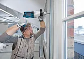 A person wearing safety equipment drills into a wall frame near a window.