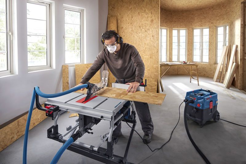 A person wearing safety equipment cuts wood on a table saw in a workshop.