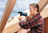 A person wearing safety equipment drills into a wooden beam at a construction site.