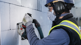 Человек, надев средства индивидуальной защиты, использует электроинструмент для прорезания пазов в бетонной стене.
