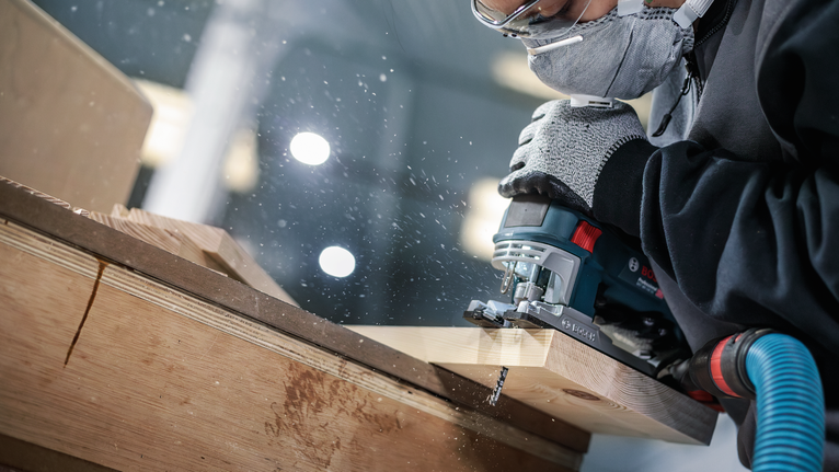 Человек, надев средства индивидуальной защиты, режет древесину электролобзиком.