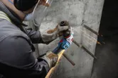 Человек в защитном снаряжении шлифует открытую арматуру на бетонной стене.