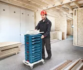 شخص يرتدي معدات السلامة ينقل صناديق الأدوات المكدسة على عربة ذات عجلات في مبنى قيد الإنشاء.