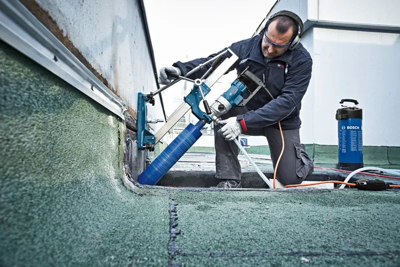 A person wearing safety equipment operates a drill stand with a core drill on a rooftop.