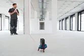 A person stands in a large room while a laser leveling tool projects a line on the floor.