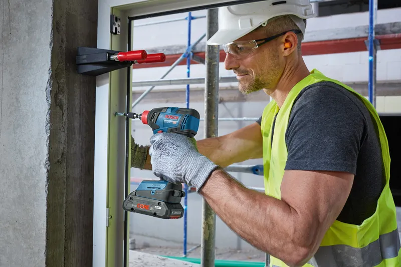 A person wearing safety equipment uses a cordless drill to fasten a window frame.