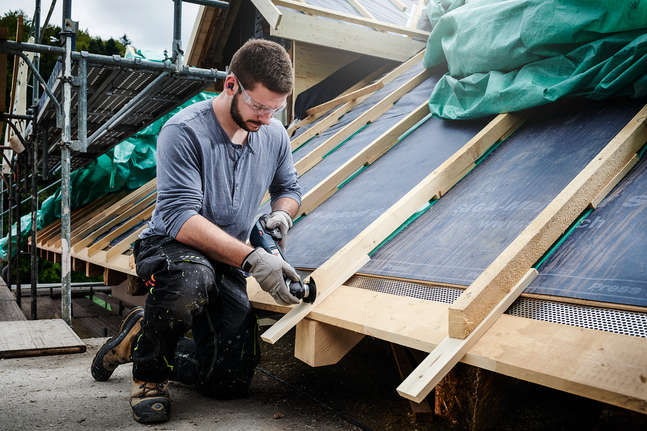 A person wearing safety equipment uses a power tool to cut wood on a rooftop construction.