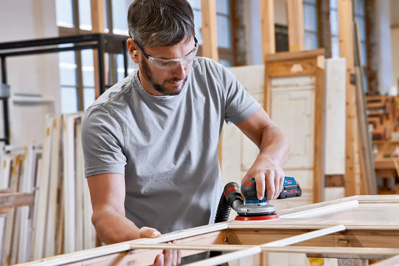 A person wearing safety equipment sands wood with a power sander in a workshop.