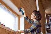 A person wearing safety equipment sands a wooden window frame with a cordless random orbit sander.