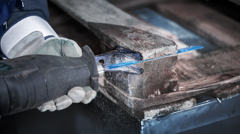 Person wearing safety equipment uses a reciprocating saw to cut through a wooden pallet.