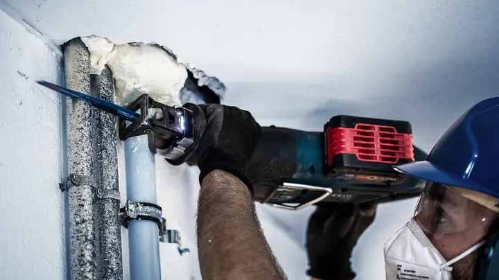 A person wearing safety equipment cuts through metal pipes using a reciprocating saw.