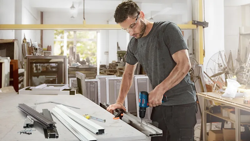 A person wearing safety equipment drills into a metal frame on a workbench.