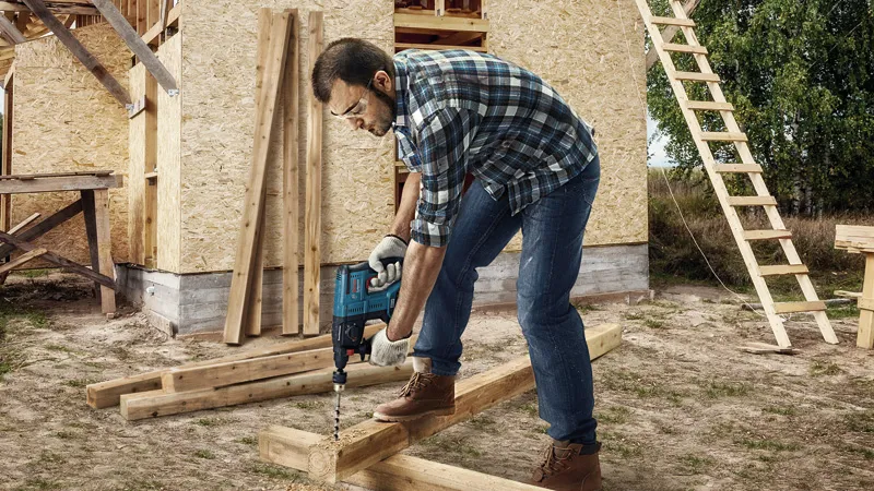 A person wearing safety equipment drills into wooden beams at a construction site.