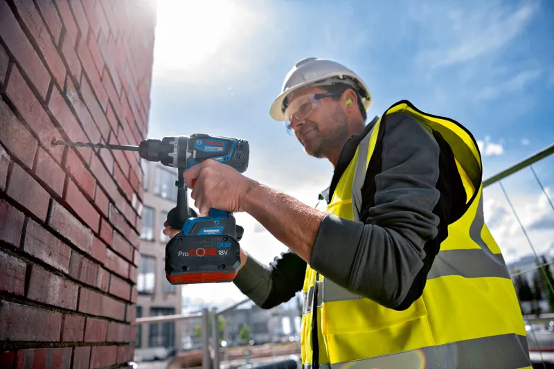 A person wearing safety equipment drills into a brick wall with a cordless combi tool.