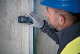 A person wearing safety equipment uses a laser measure against a concrete wall.