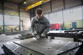 Person wearing safety equipment grinds a large metal sheet in a factory workspace.