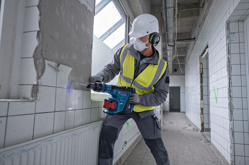 A person wearing safety equipment uses a cordless rotary hammer to break tiled wall.