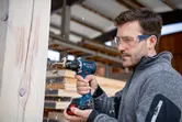 Person wearing safety equipment drills into a wooden beam in a workshop.