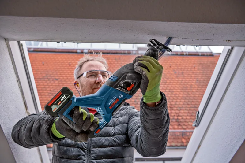 A person wearing safety equipment uses a cordless reciprocating saw to cut a window frame.