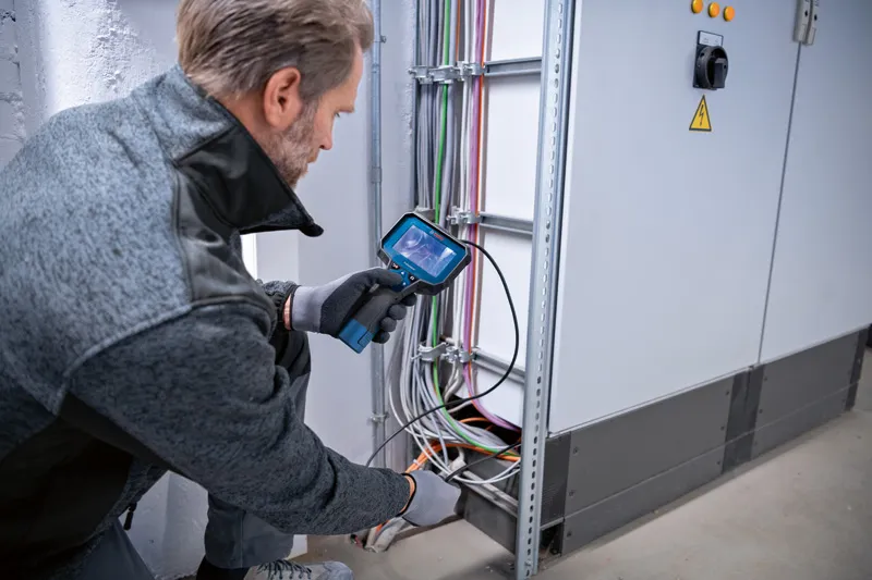 A person wearing safety equipment inspects electrical wiring using an inspection camera.