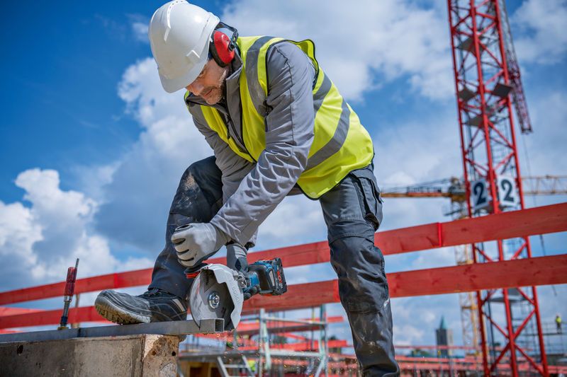 A person wearing safety equipment uses a cordless angle grinder to cut metal at a construction site.