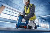 A person wearing safety equipment operates a blue core drill on a concrete floor.